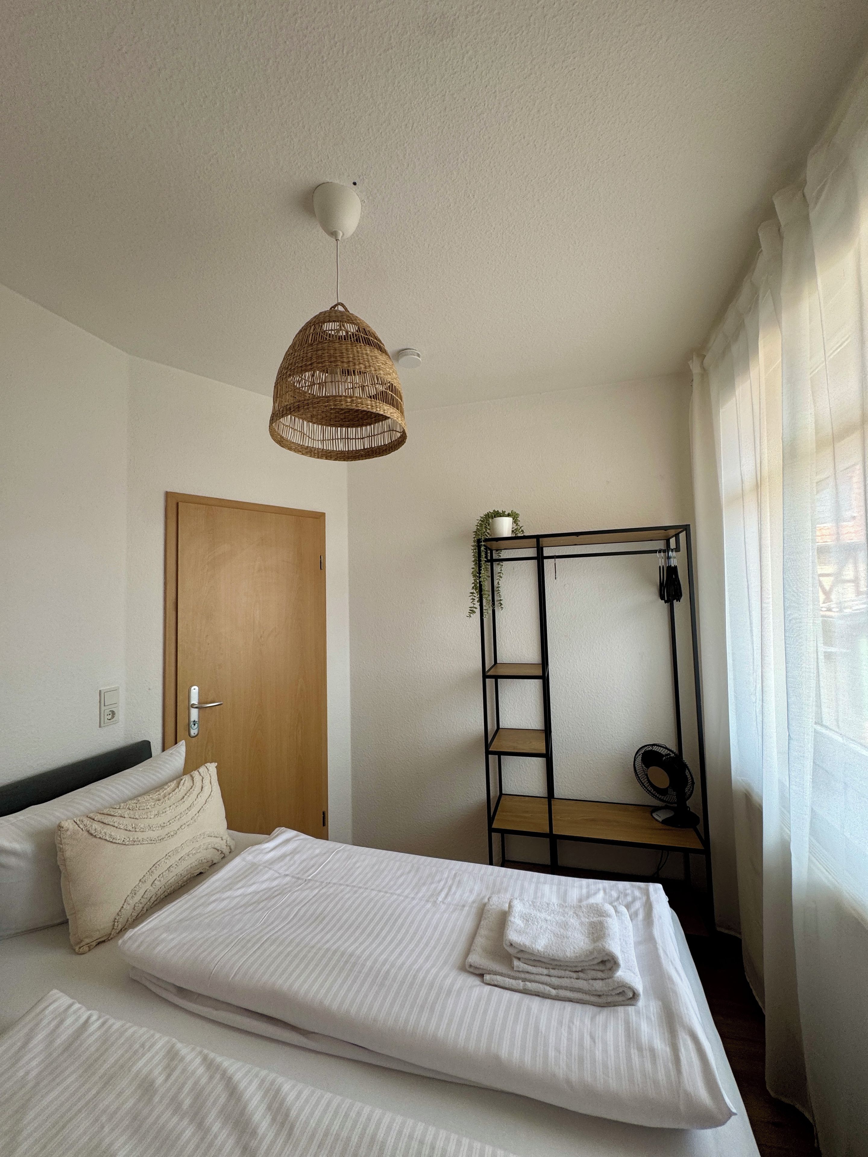 A bedroom with a bed, a hanging light fixture, a wooden door, a shelf with a plant, and a window with curtains.