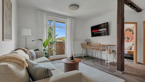 A modern hotel room with a sofa, dining area, and a TV displaying the hotel name. - REGIOHOTEL am Nicolaiplatz Wernigerode (Wernigerode)