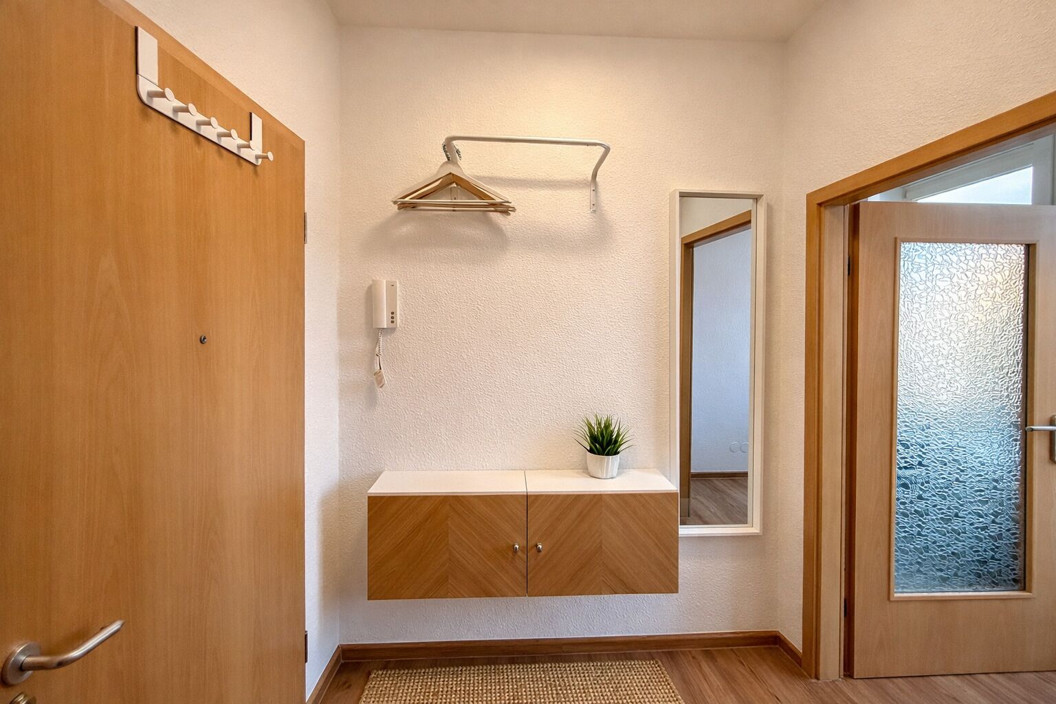 Un pasillo con una puerta de madera, un perchero, una planta pequeña, un espejo y una puerta de cristal satinado.