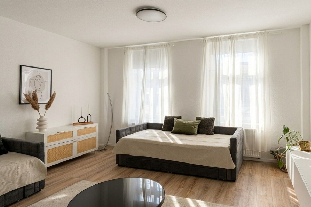 A modern bedroom with a large bed, a bedside table, a TV, and a window with sheer curtains.