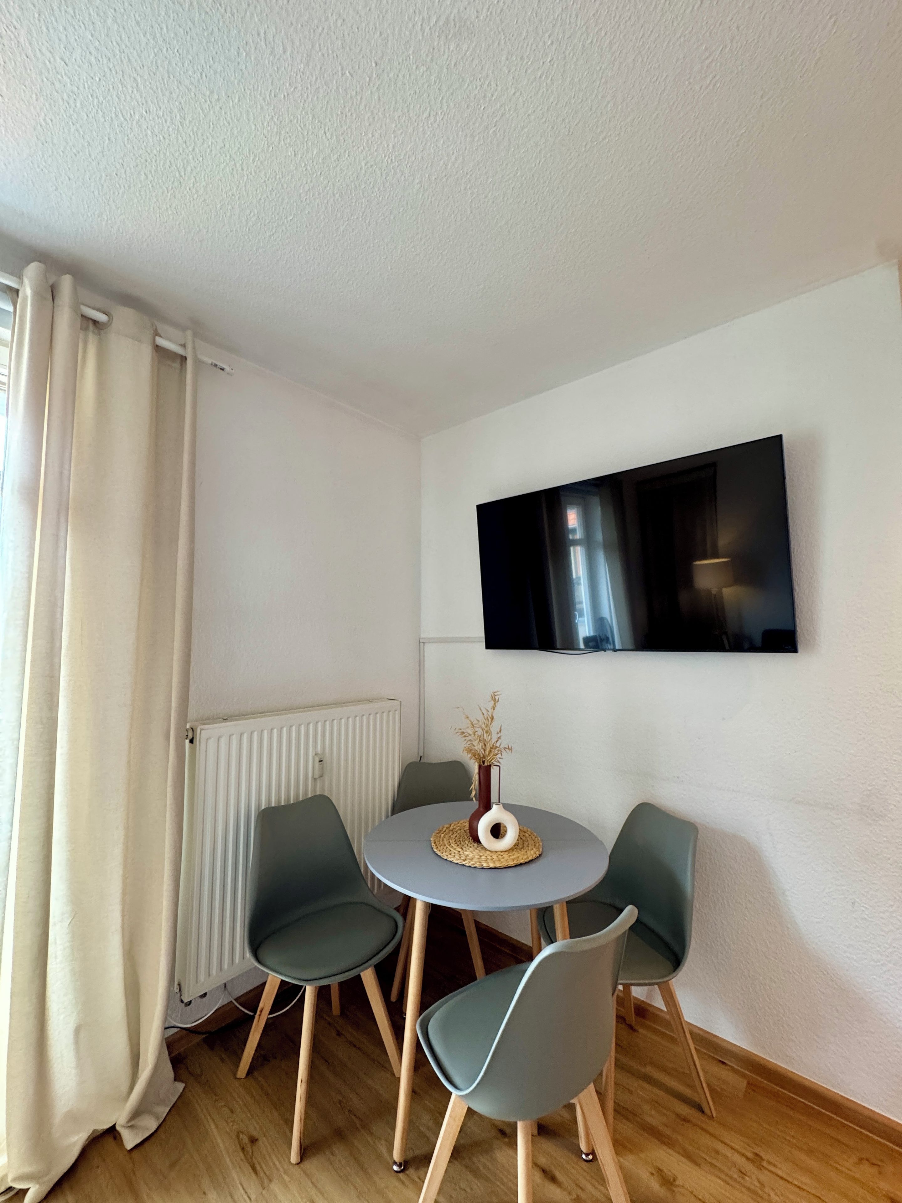 Een kleine kamer met een ronde tafel, twee stoelen, een flatscreen televisie aan de muur en een radiator.