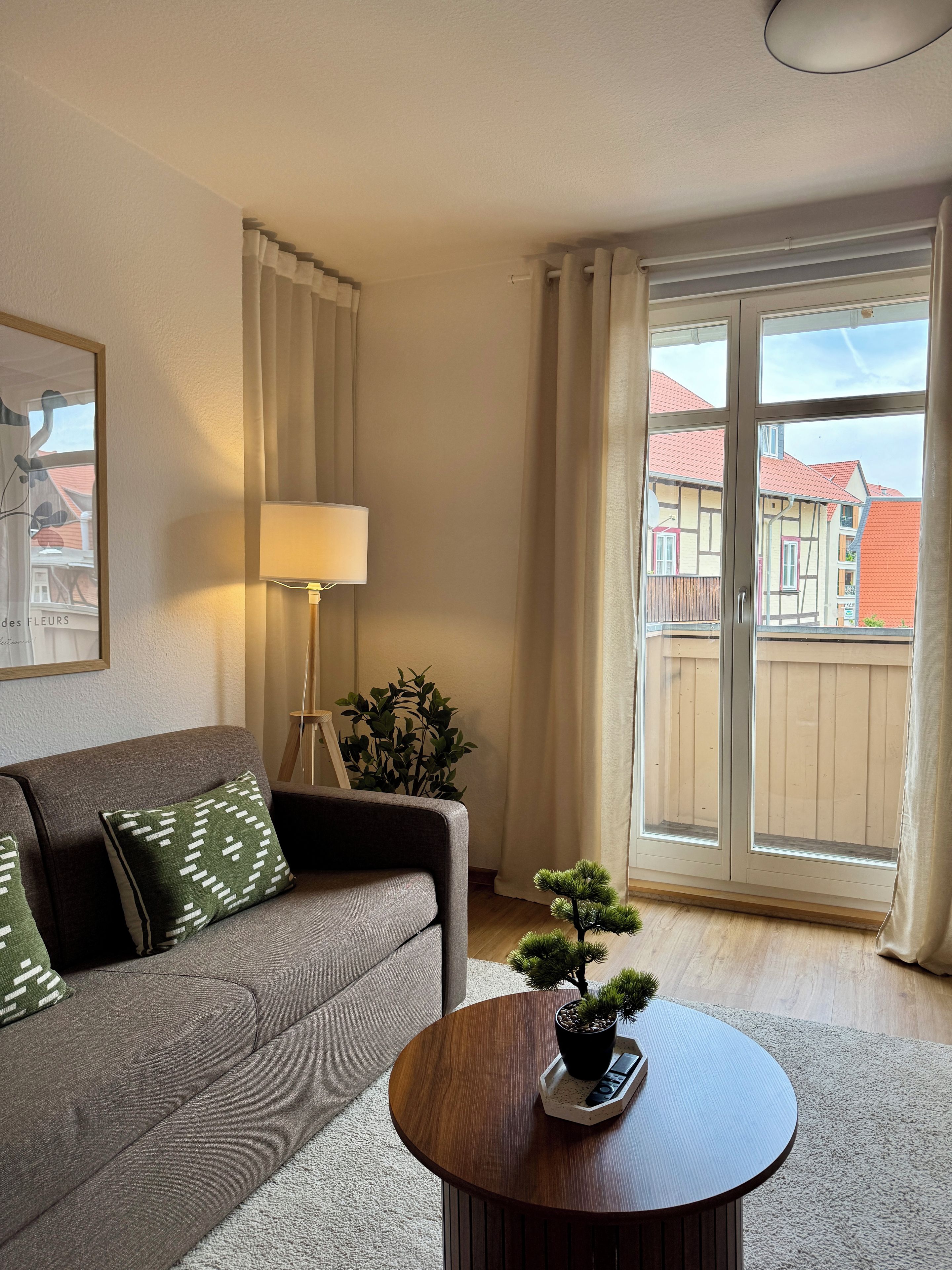 En stue med sofa, lampe, lille bord med plante og udsigt til bygninger gennem et vindue.