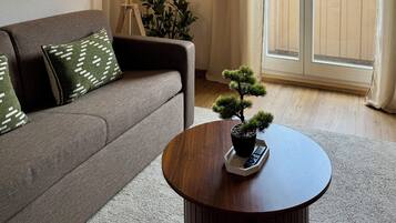 En stue med sofa, lampe, lille bord med plante og udsigt til bygninger gennem et vindue.