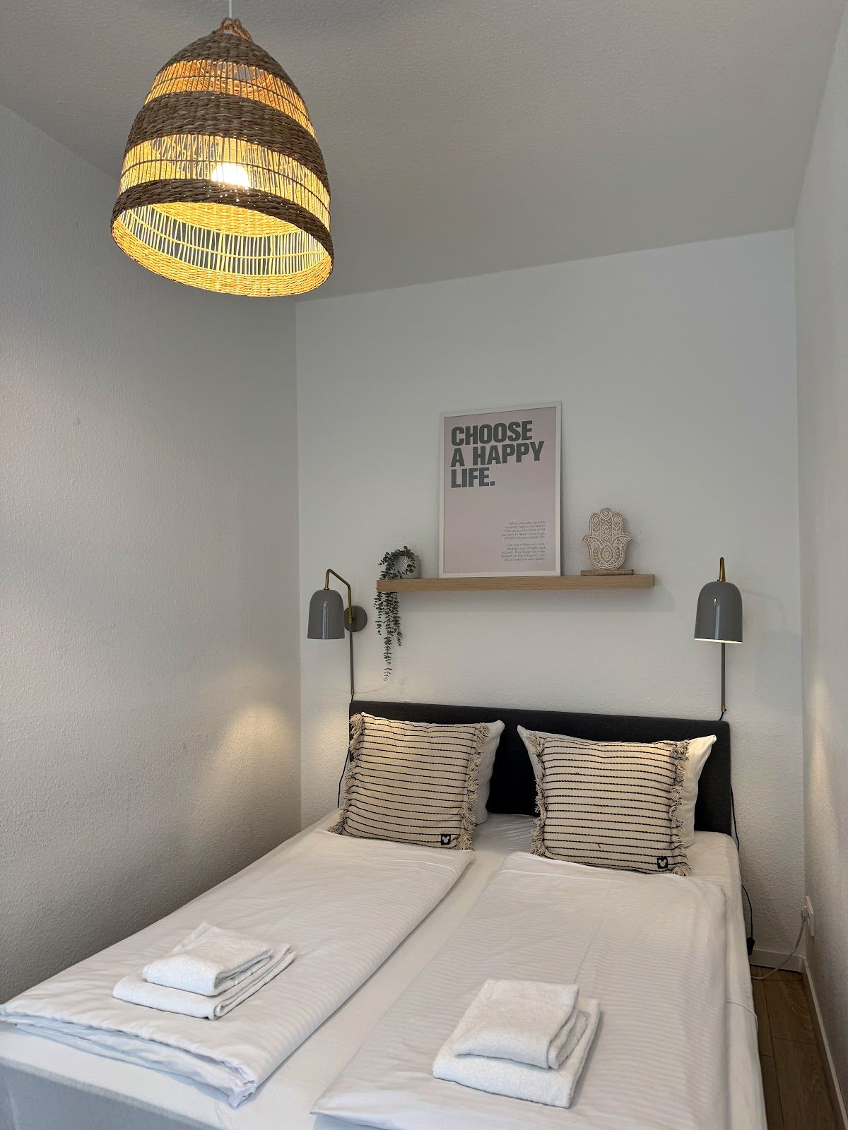 Ein Schlafzimmer mit einem Bett, zwei Nachttischlampen, einem an der Wand befestigten Regal mit einer Pflanze und einer Pendelleuchte.