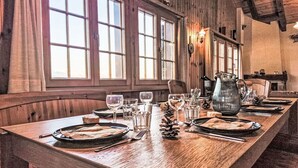 A long wooden dining table set with plates, glasses, and cutlery, flanked by windows and a stone fireplace.
