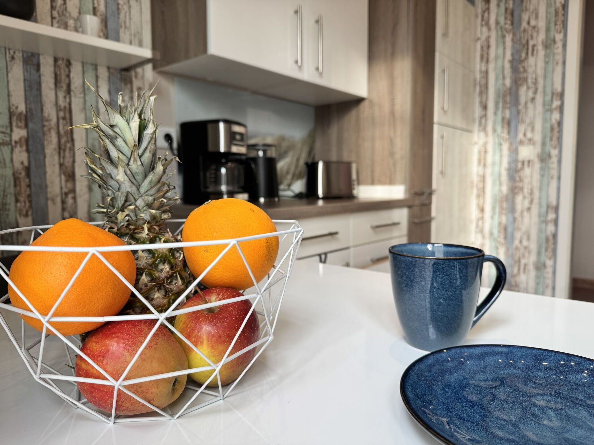 A kitchen with a fruit bowl containing apples, oranges, and a pineapple, a blue mug, and a blue plate on the counter.