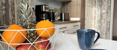 A kitchen with a fruit bowl containing apples, oranges, and a pineapple, a blue mug, and a blue plate on the counter.
