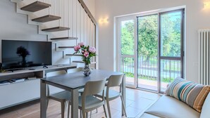 A modern living room with a staircase, a flat-screen TV, a dining table with chairs, a sofa, and a large window with a view of greenery. - Lovely apartment for 4 guests with A/C, pool, WIFI, TV and balcony (Domaso)