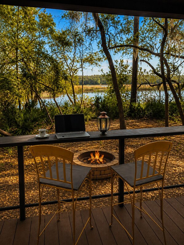 A deck with a fire pit, two chairs, a laptop, and a cup on a table overlooking a lake.