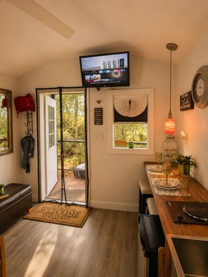 A modern kitchen with a television mounted above the counter, a dining area with a table set for two, and a large window with a view of the outdoors. - Romantic Hideaway Cabin • Hammock • Fire Pit • Free Parking • Pet-Friendly.  (Hawthorne)