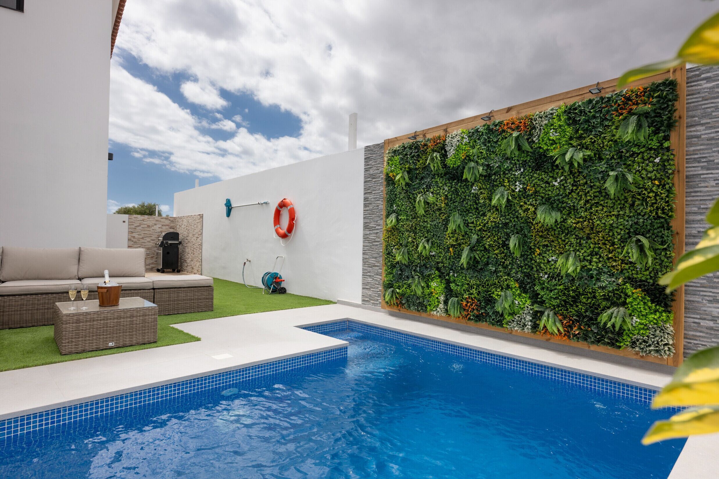 Una piscina con borde de azulejos, zona de césped artificial verde, sofá de mimbre y una pared con jardín vertical.