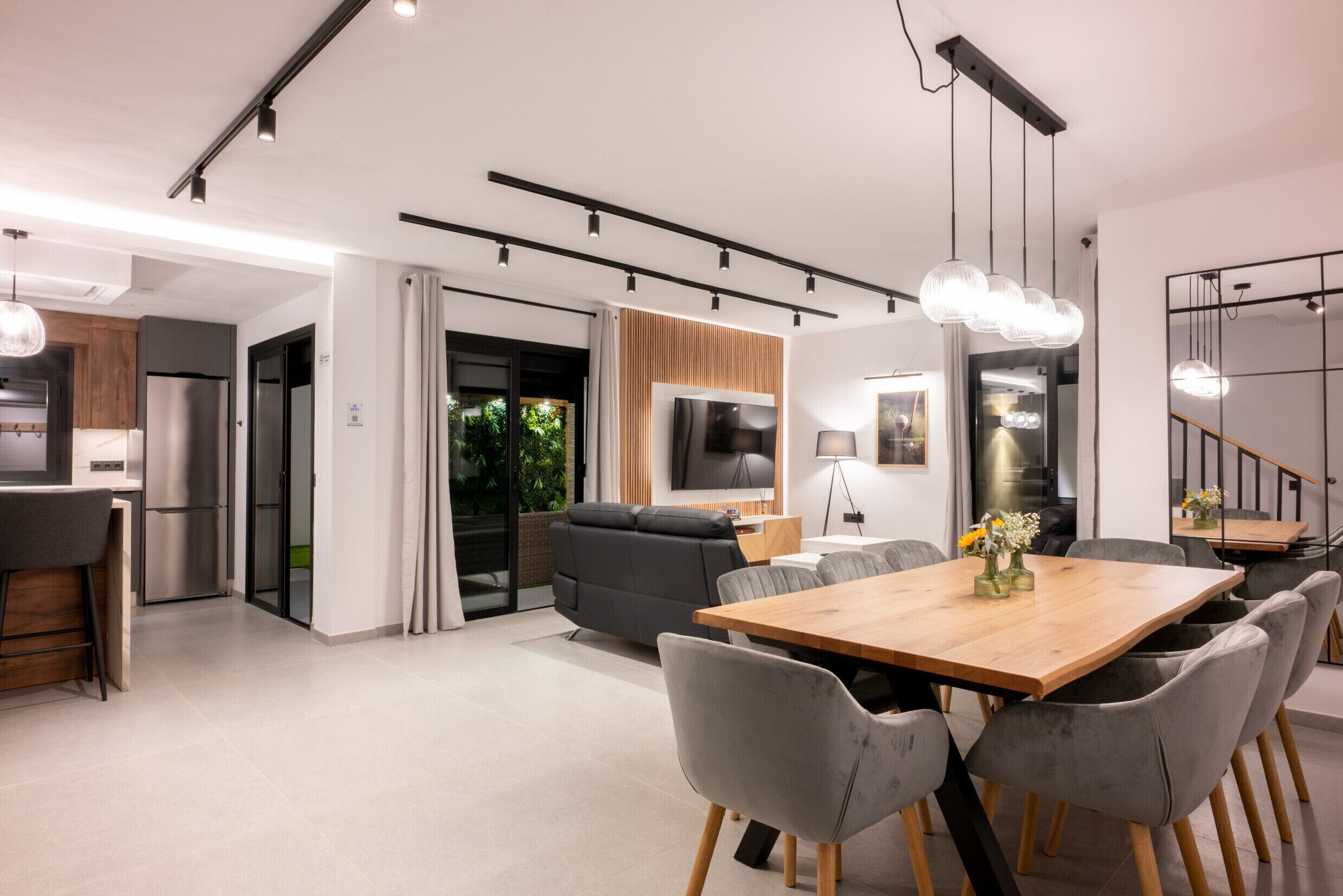 Una cocina moderna con horno empotrado, una zona de comedor con mesa de madera y sillas grises, y un área de estar con sofá y televisor.