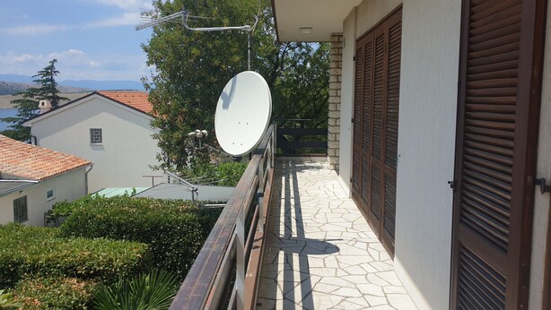 Un balcon doté d’une antenne parabolique, d’un sol carrelé en pierre et offrant une vue sur les bâtiments et les arbres environnants.