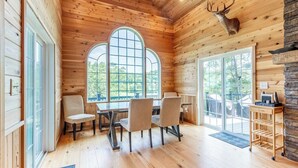 Interior de una cabaña de madera con una mesa de comedor y sillas, ventanas arqueadas amplias y una cabeza de ciervo montada.