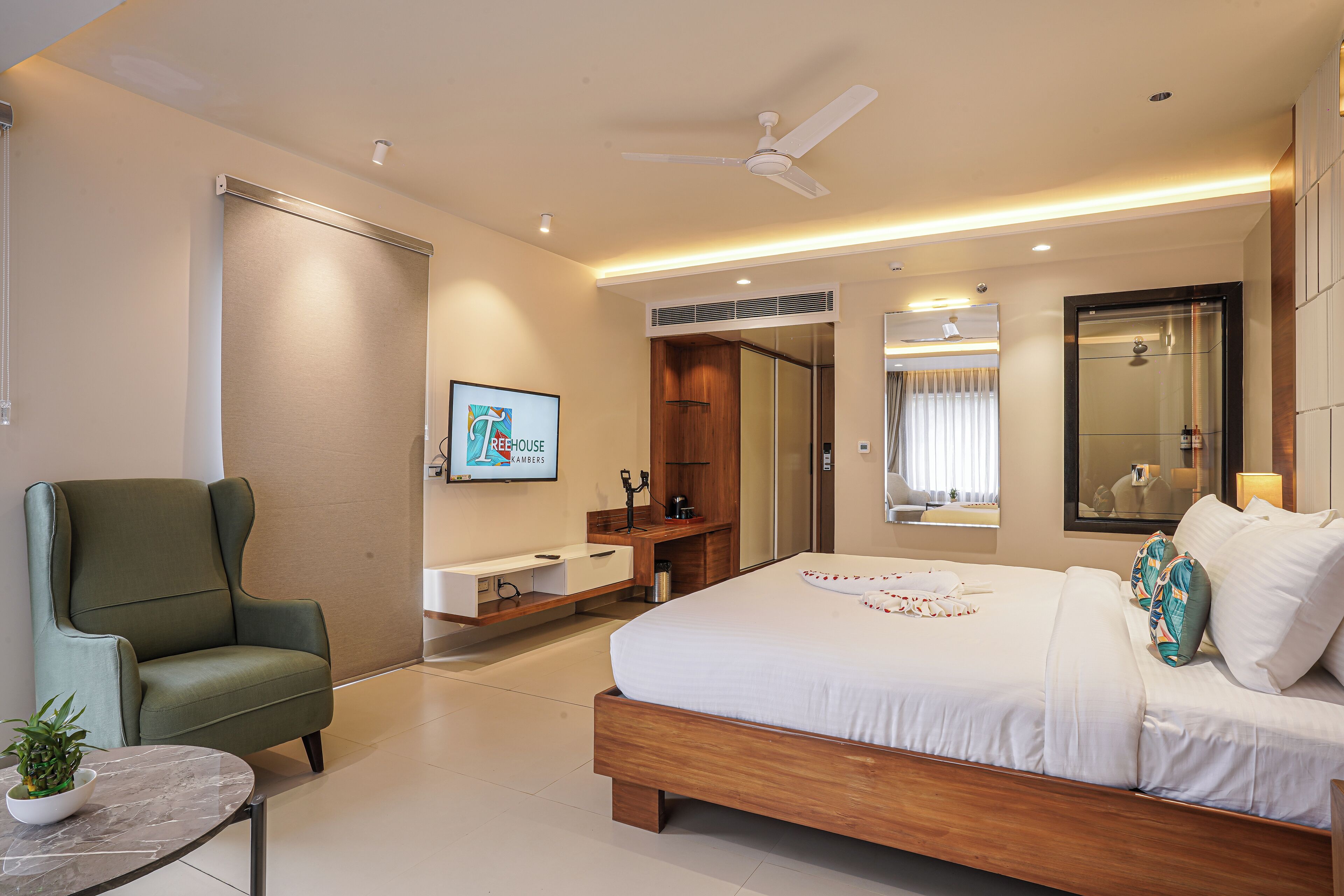 A modern hotel room with a large bed, a green armchair, a small table with a plant, a bathroom with a vanity, and a ceiling fan.