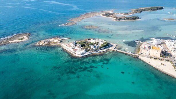 An aerial view of a coastal area with clear turquoise waters, several small islands, and a main island with buildings and a paved area. - Villetta Sogno Di Mare In Porto Cesareo (Porto Cesareo)