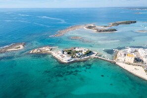 Ett flygbildsbild av ett kustområde med klart turkosfärgat vatten, flera små öar och en större ö med byggnader och ett asfalterat område.