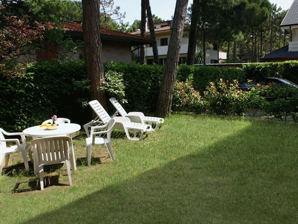 A grassy outdoor area with white lounge chairs and a table, surrounded by trees and a building in the background.