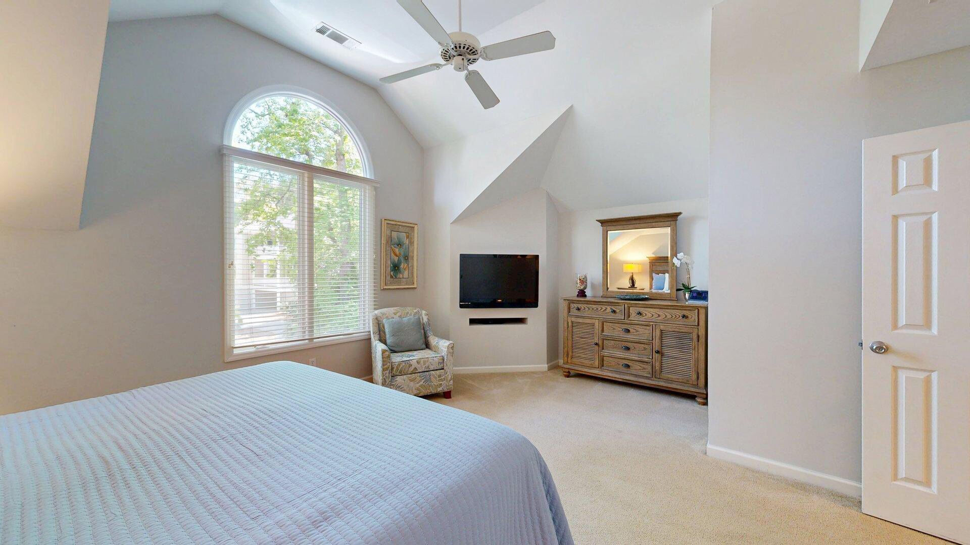 Une chambre avec un grand lit, un ventilateur de plafond, une télévision, une commode et une fenêtre avec des stores.