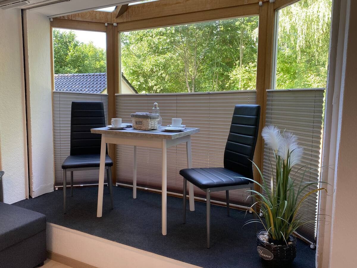 Un patio cubierto con una mesa y dos sillas, una planta en maceta y vistas a árboles a través de las persianas.