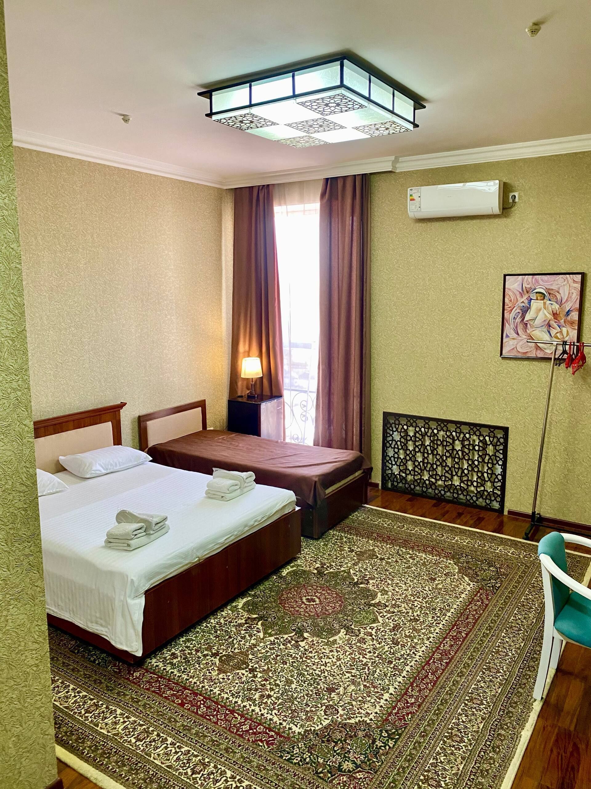 A hotel room with two beds, a large patterned rug, an air conditioning unit, and a window with curtains.