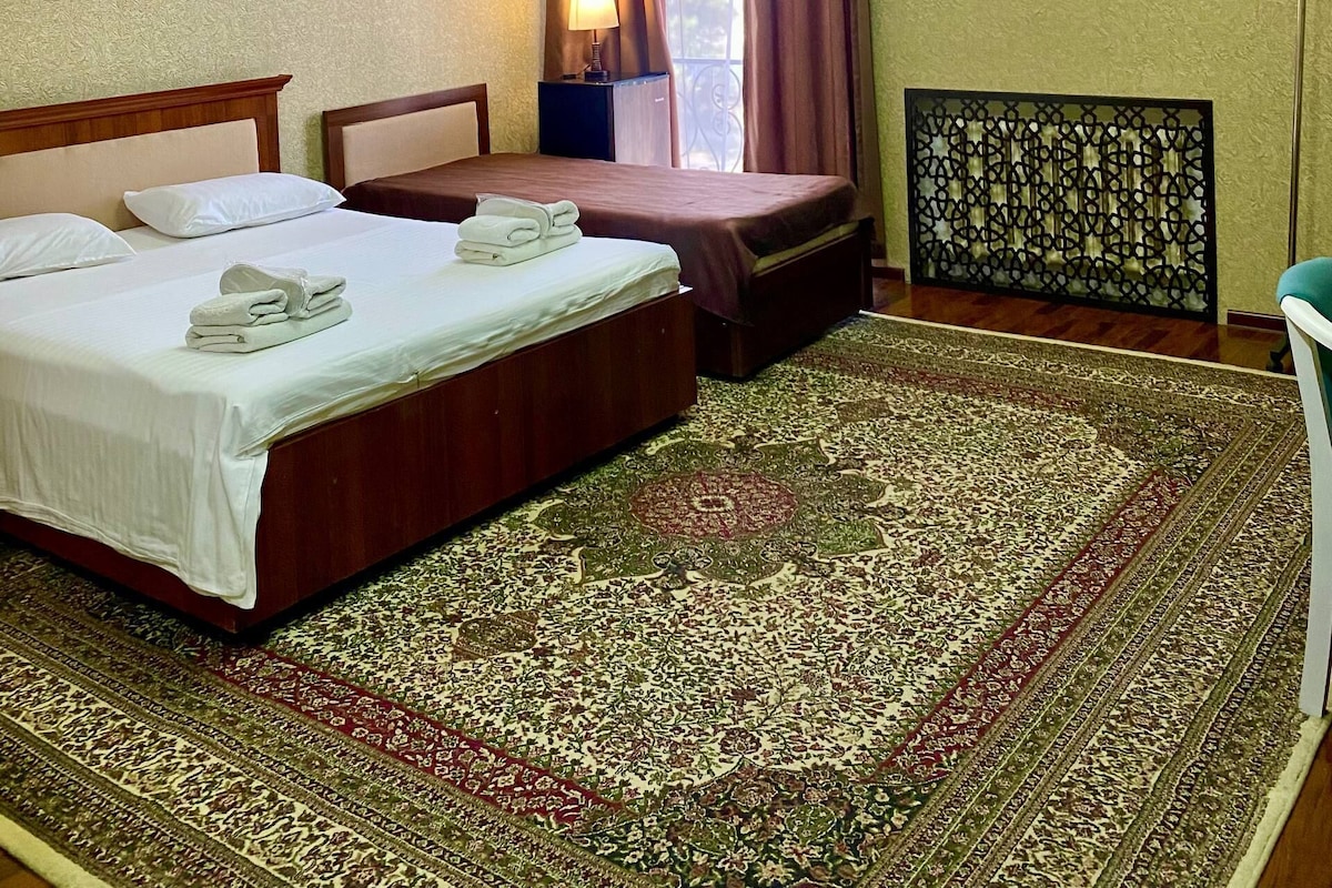 A hotel room with two beds, a large patterned rug, a ceiling light fixture, and an air conditioning unit.