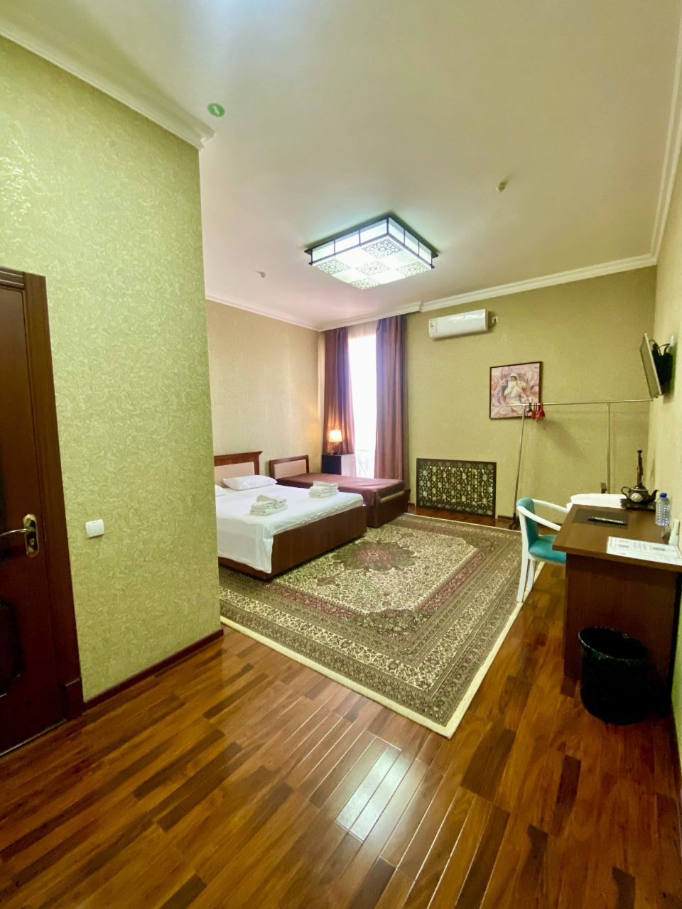 A hotel room with a wooden floor, a bed with a headboard, a desk with a chair, a radiator, and a window with curtains.