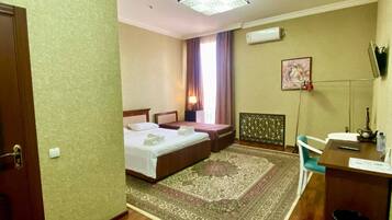 A hotel room with a wooden floor, a bed with a headboard, a desk with a chair, a radiator, and a window with curtains.