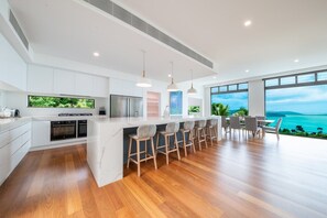 A modern kitchen with a central island, bar stools, and a view of the sea. - CoralAir Whitsundays (Airlie Beach)
