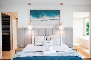A modern bedroom with a large bed, a painting of a beach scene, and a view of a bathroom with a bathtub. - CoralAir Whitsundays (Airlie Beach)