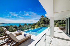 A modern outdoor pool area with lounge chairs, a glass balustrade, and a view of the sea. - CoralAir Whitsundays (Airlie Beach)