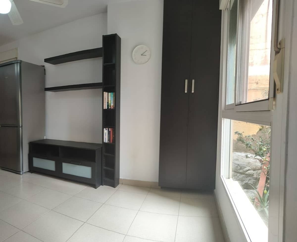 A room with a dark bookshelf, a clock on the wall, and a window with a view of the outside.