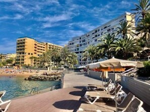 A beachfront hotel with a poolside area, palm trees, and a beach in the background.