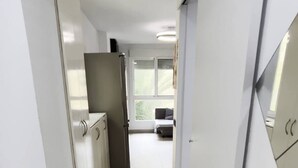 Un couloir étroit doté d’un parquet en bois de couleur claire, de murs blancs et offrant une vue sur un espace de vie comprenant un canapé et une fenêtre.