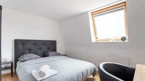 Un dormitorio moderno con una cama grande, una mesita de noche pequeña, una ventana con persianas, una silla negra y suelo de madera.