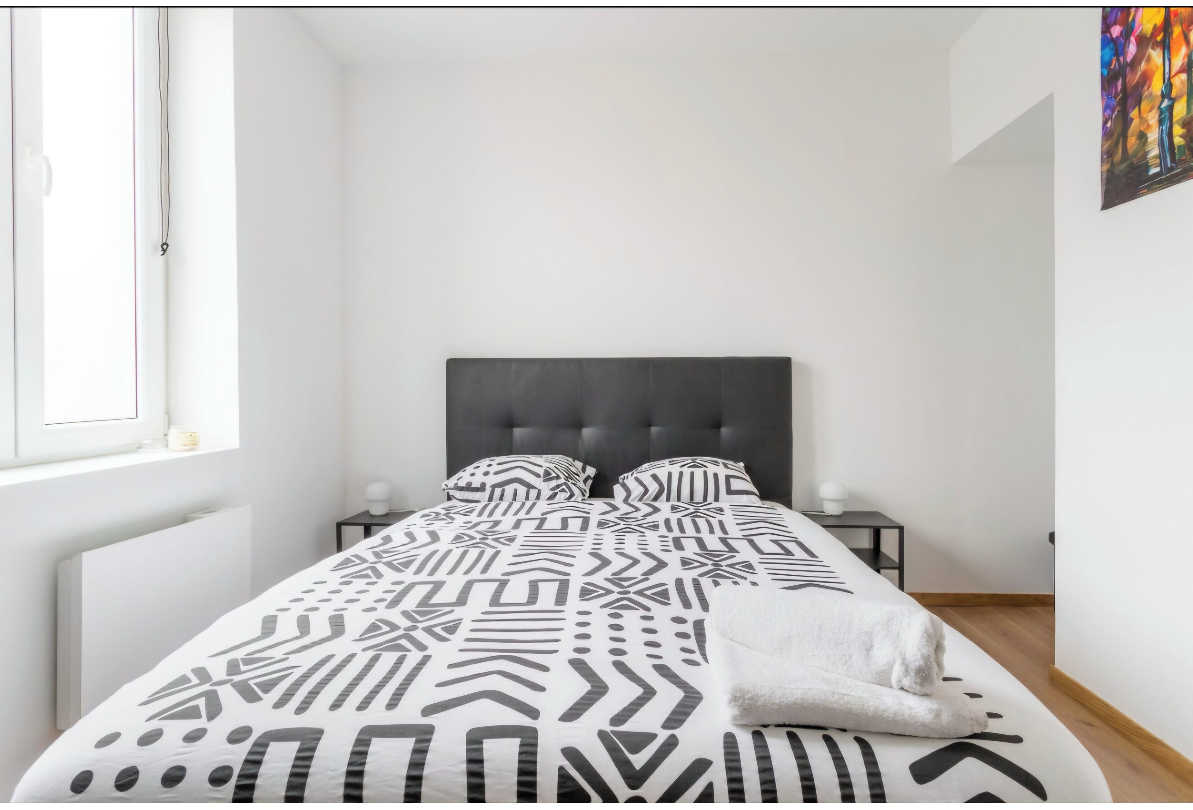 Une chambre à coucher moderne avec un grand lit, une tête de lit sombre et une fenêtre laissant entrer la lumière naturelle.