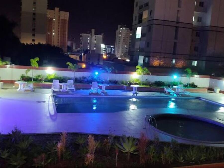 Un área de piscina en la azotea con sillones de descanso y un jacuzzi, rodeada de plantas en macetas e iluminada con luces ambientales.