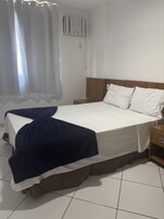 Cama bem arrumada com lençóis brancos e manta azul, mesa de cabeceira de madeira com abajur, e janela com cortinas leves.