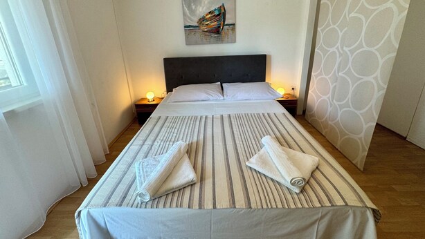 Een net opgemaakt bed met gestreepte beddengoed en twee handdoeken, een schilderij aan de muur en een kamerbreed gordijn met patroon.