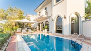 Ein Schwimmbecken mit weißem Beckenrand, einer überdachten Terrasse und einem zweigeschossigen Gebäude mit Bogenfenstern und einem Balkon.