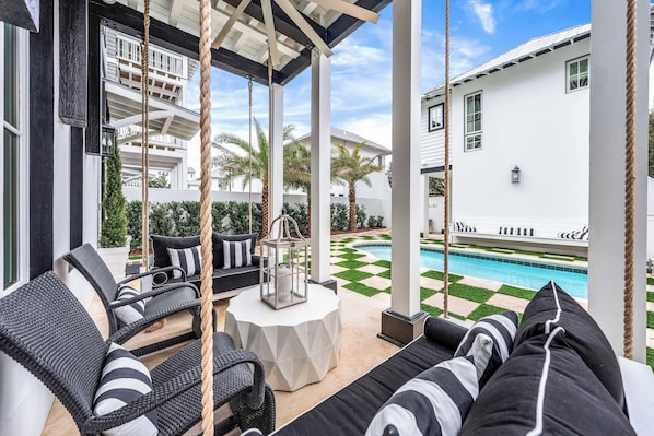 A covered outdoor seating area with wicker furniture, a pool, and a white building in the background.