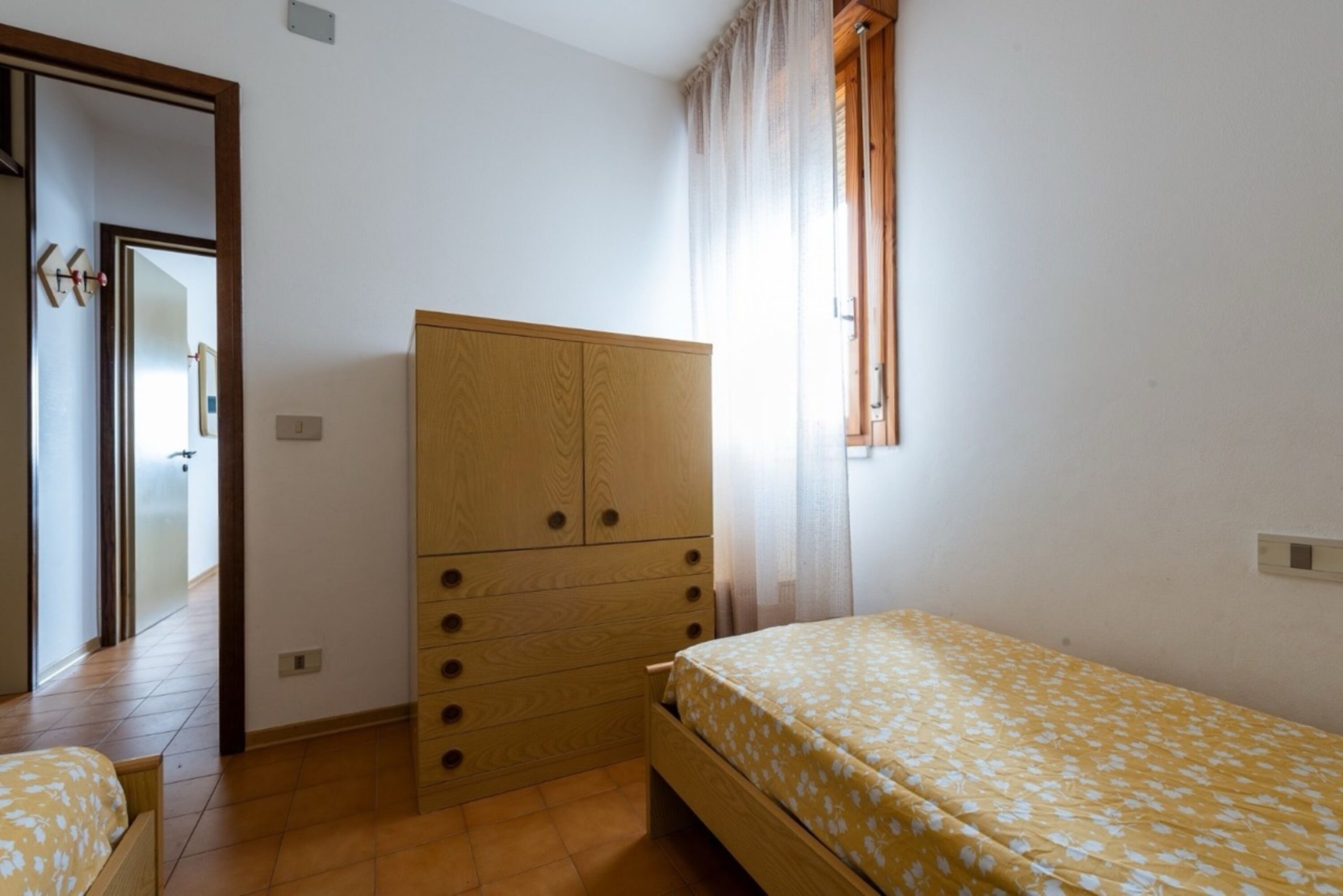 A bedroom with a wooden wardrobe, a bed with a floral bedspread, and a window with curtains.