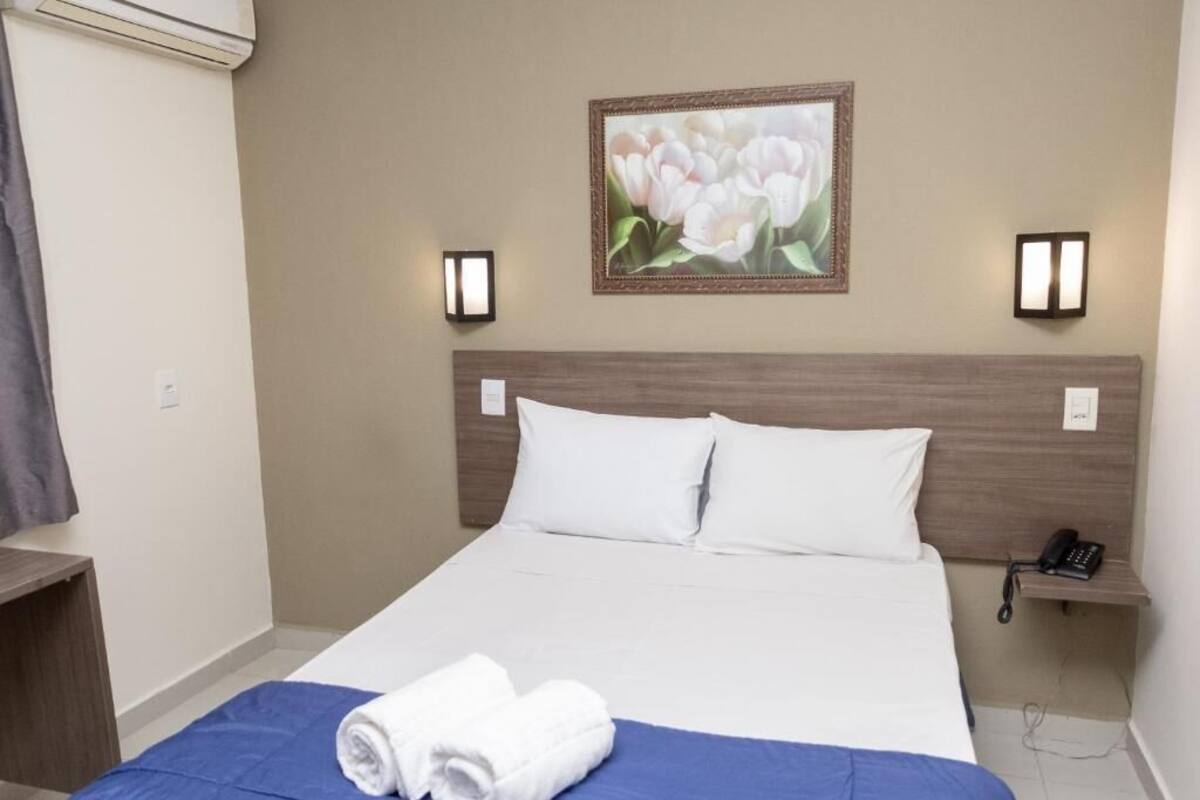 A hotel room with a bed, a wall-mounted painting, bedside lamps, a phone, and a folded towel on the bed.