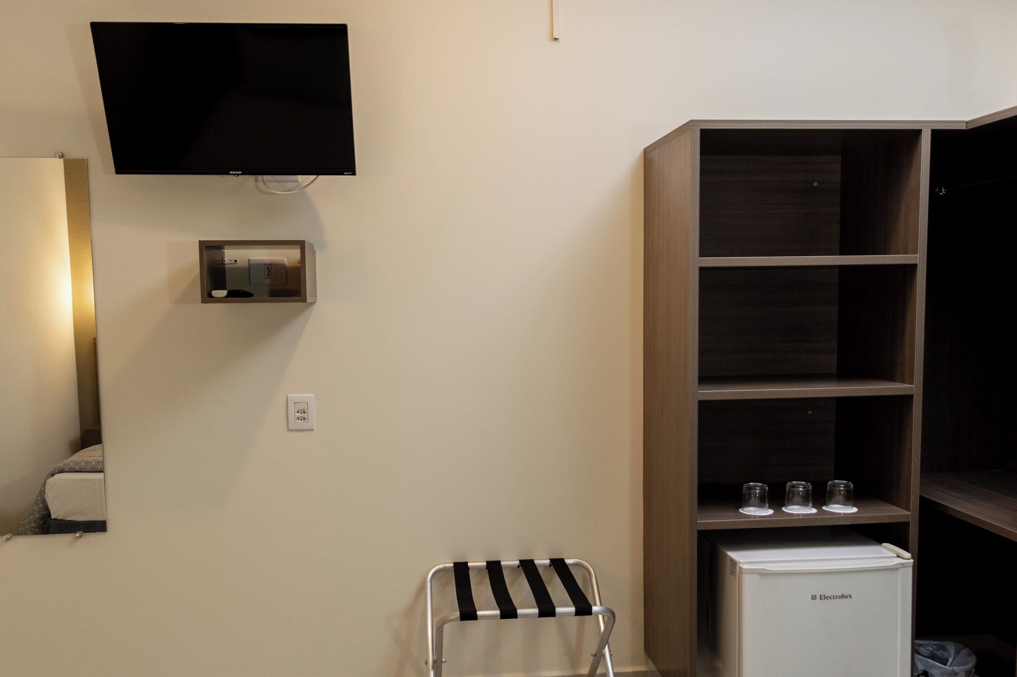 Habitación compacta con un televisor de pantalla plana montado en la pared, un pequeño panel de control debajo, un refrigerador blanco y una silla de metal con un cojín a rayas negras y blancas.
