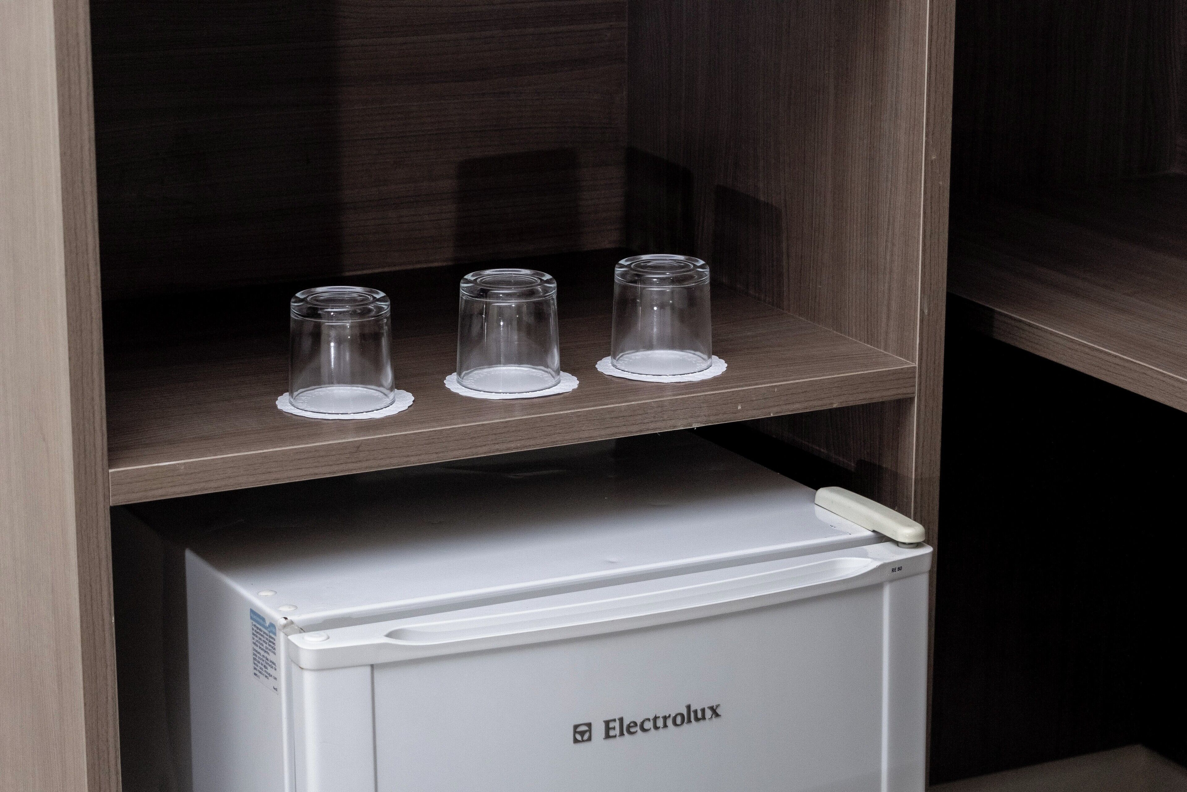 Uma geladeira branca Electrolux com painel de controle, posicionada sob uma prateleira de madeira com três copos em bases.
