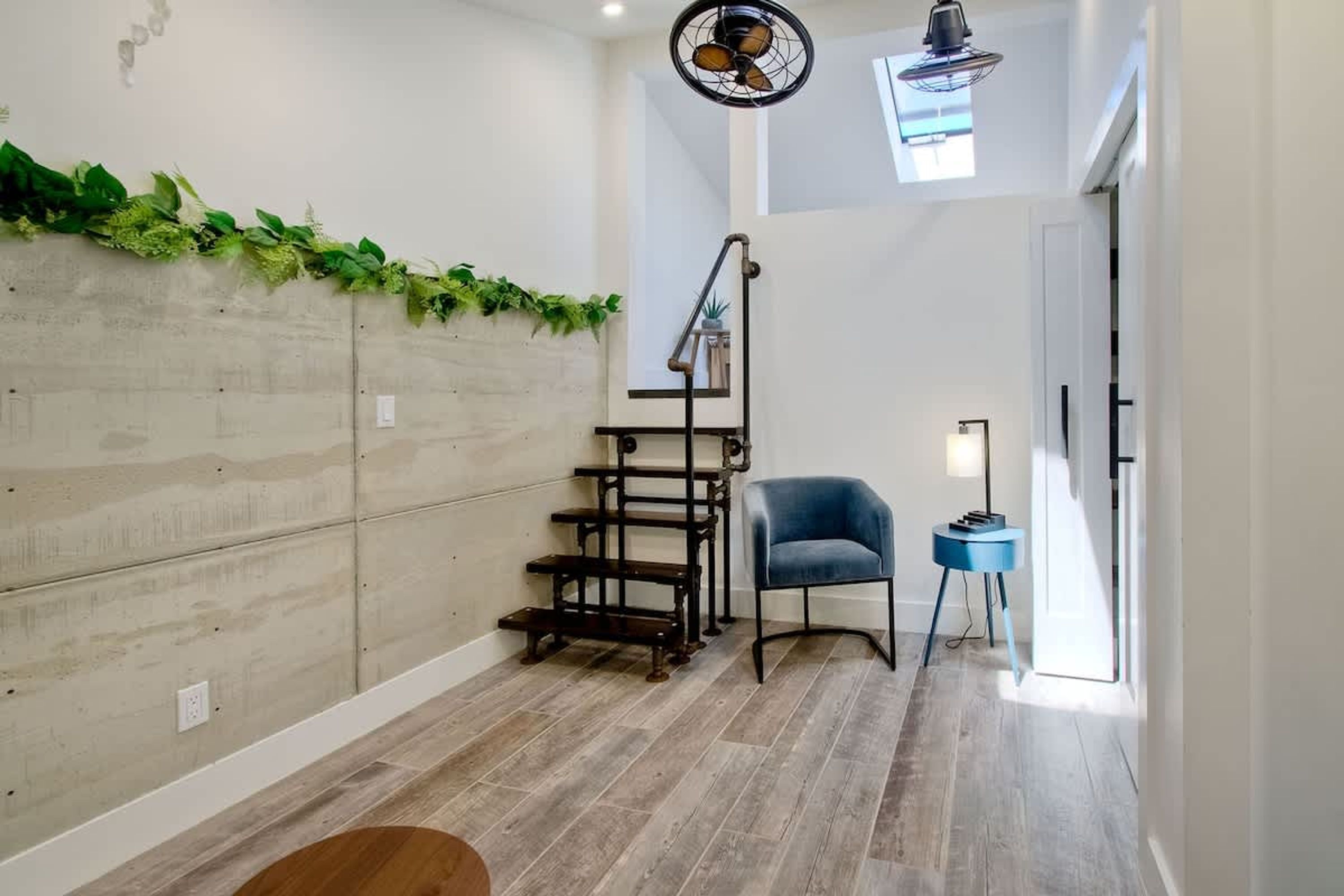 Un intérieur moderne avec un plancher en bois, une chaise bleue, une petite table et un escalier décoré d’une guirlande de plantes.