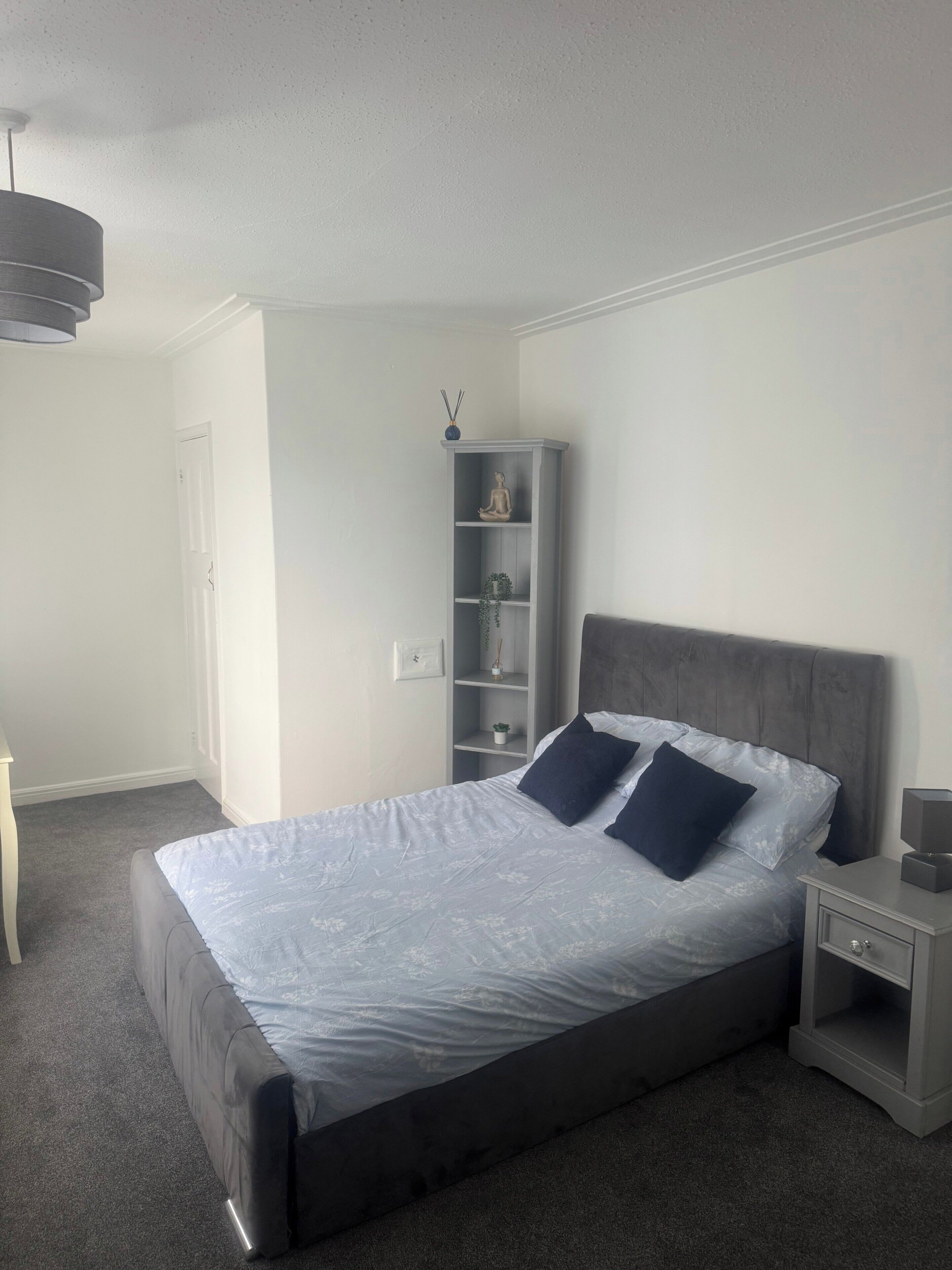 A bedroom with a bed, a nightstand, a shelf with decorative items, and a door.