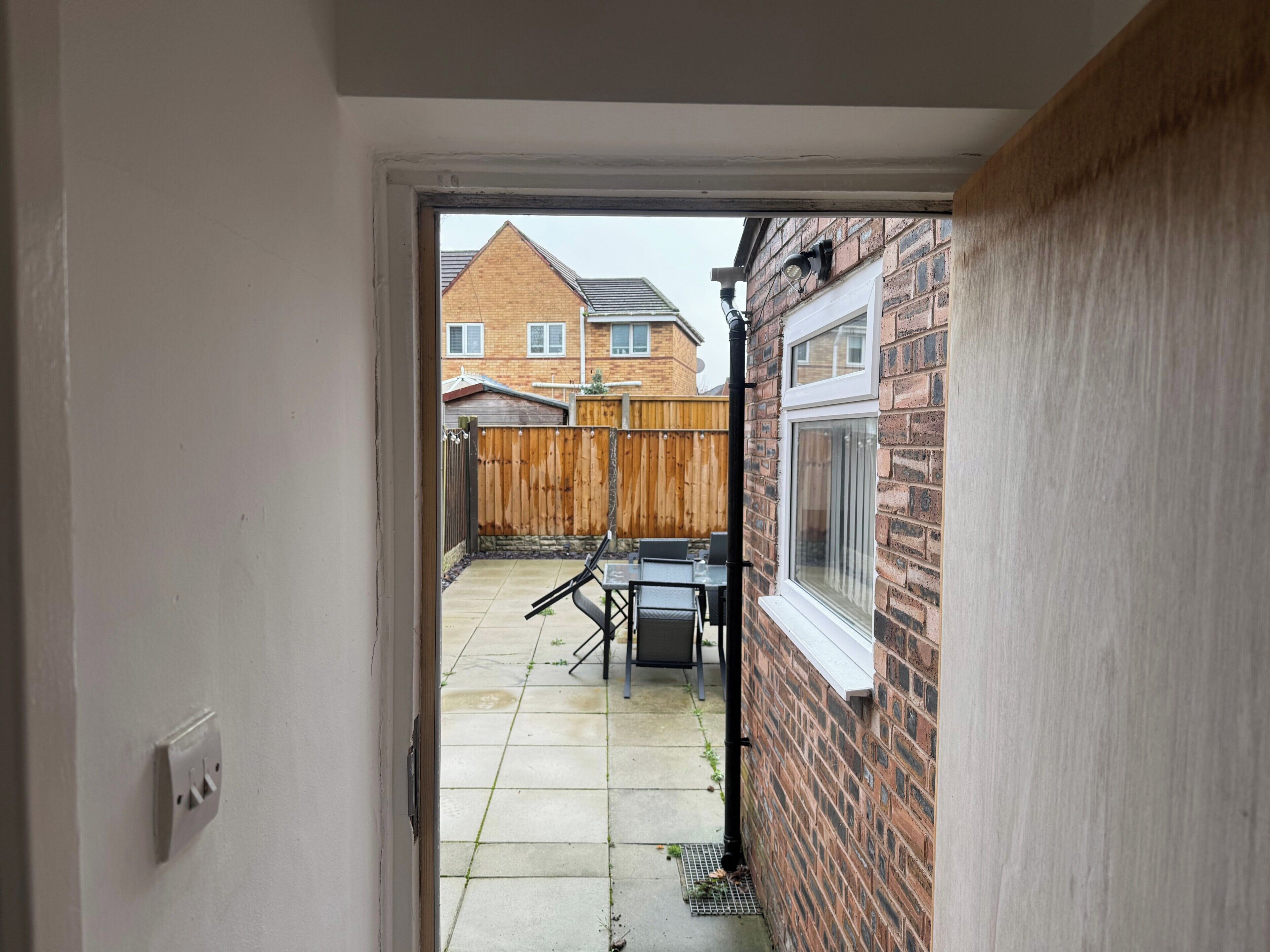 A view through a doorway into a backyard with a patio table and chairs, a wooden fence, and a brick wall with a window.