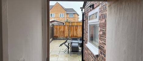 A view through a doorway into a backyard with a patio table and chairs, a wooden fence, and a brick wall with a window.