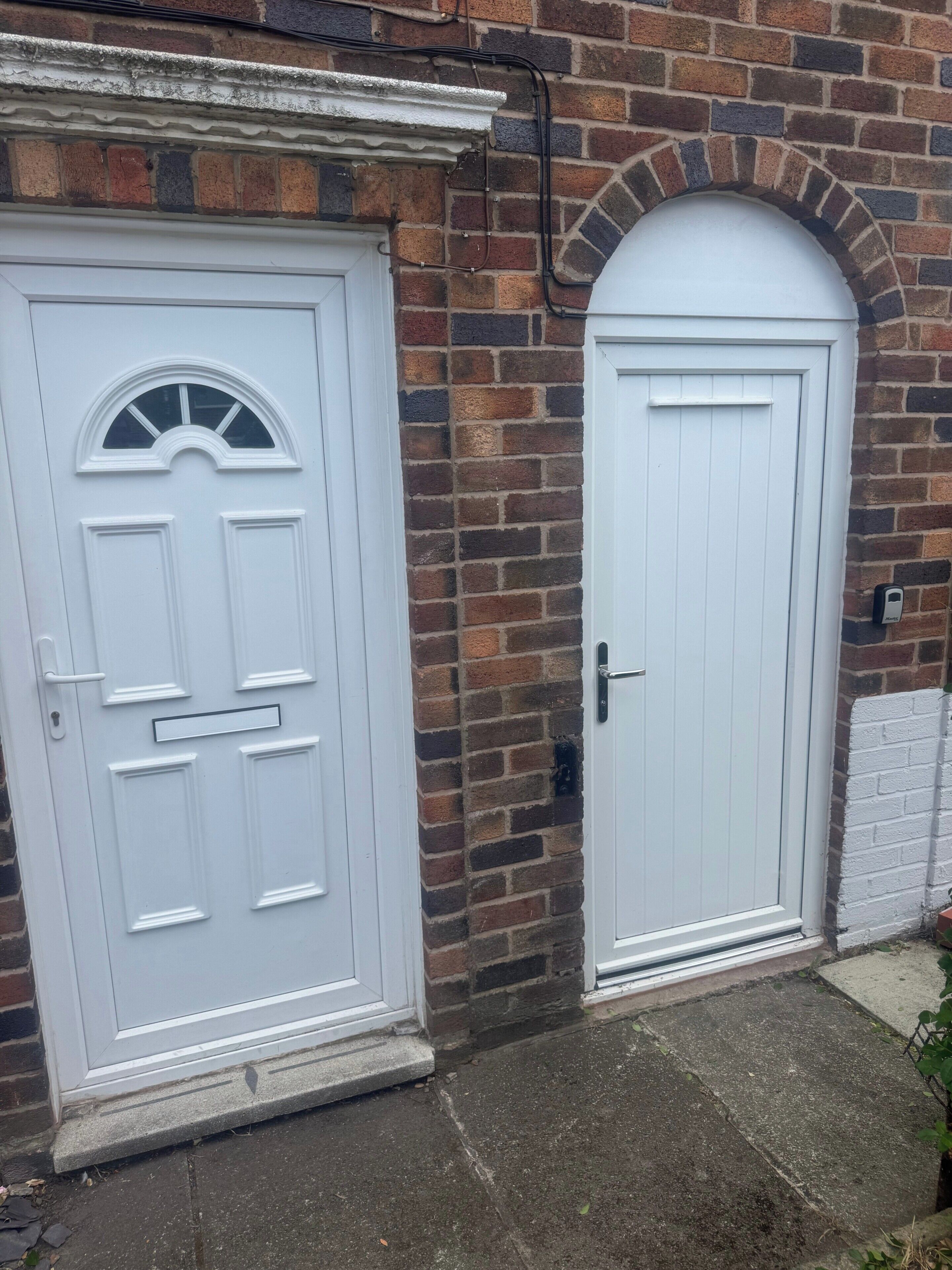 Two white doors with different designs, one with a semi-circular window and the other with a plain design, set against a brick wall with a white trim.